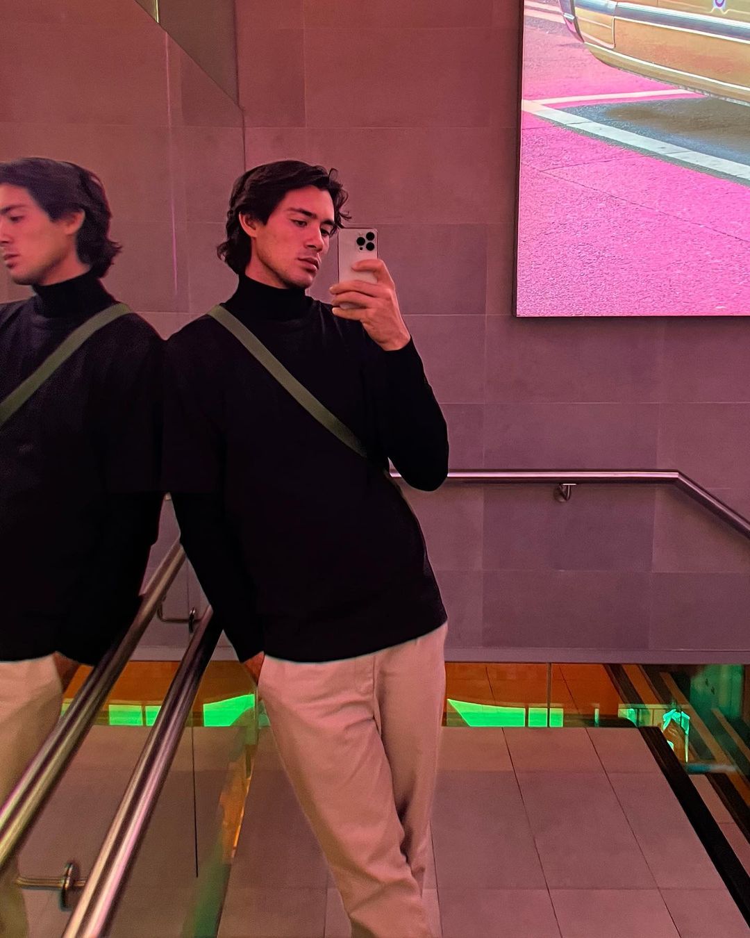 Man with dark hair in black turtleneck and khaki pants takes selfie in pink and purple lit interior with mirror reflection.