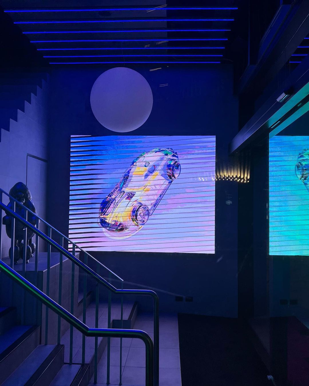 A modern interior showcases a digital display featuring a translucent car, set against a blue-lit backdrop and illuminated staircase.