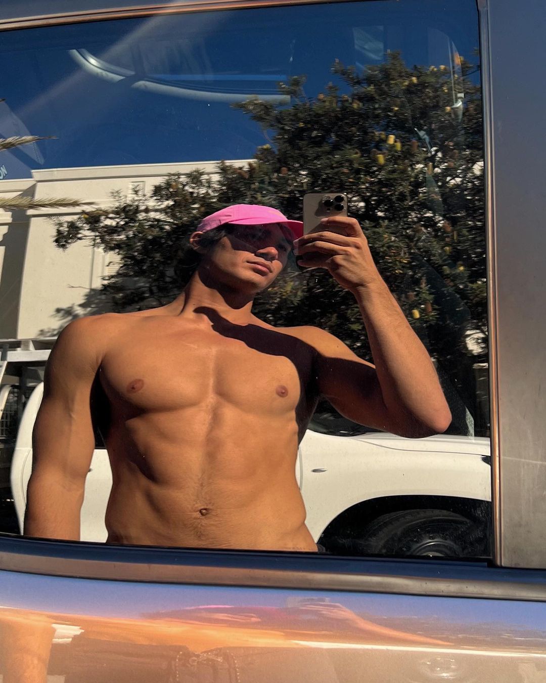 Shirtless man with pink baseball cap takes a selfie in a car with a tree and building visible in the reflection outdoors.