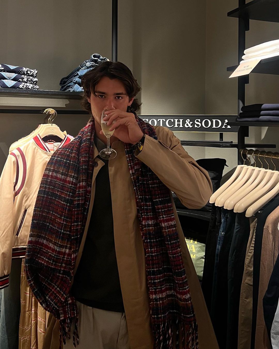 Man in overcoat and scarf holds champagne glass inside clothing store with shelves of merchandise and 'Scotch & Soda' signage.
