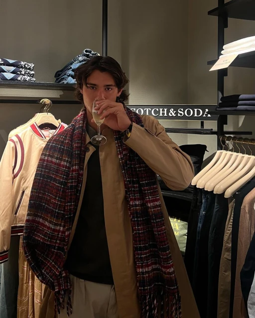 Man in overcoat and scarf holds champagne glass inside clothing store with shelves of merchandise and 'Scotch & Soda' signage.