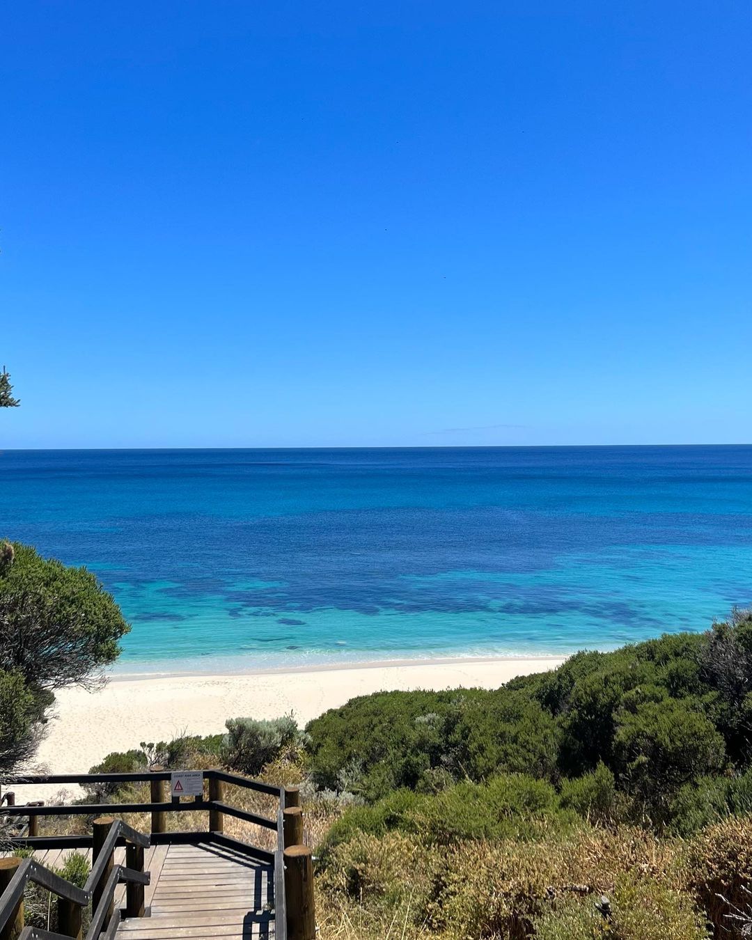 Coastal view with clear blue sky, turquoise ocean water, white sand beach, lush green vegetation, and wooden stairs leading down.
