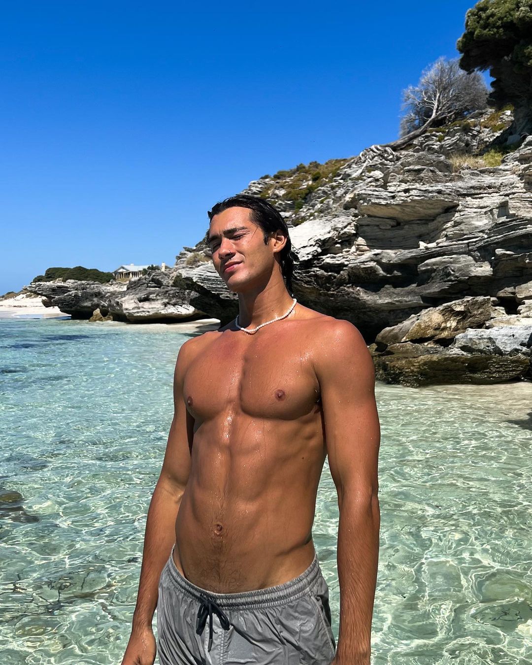 Adult man with athletic physique stands in shallow, clear water at the beach on a sunny day with a rocky coastline in the background.