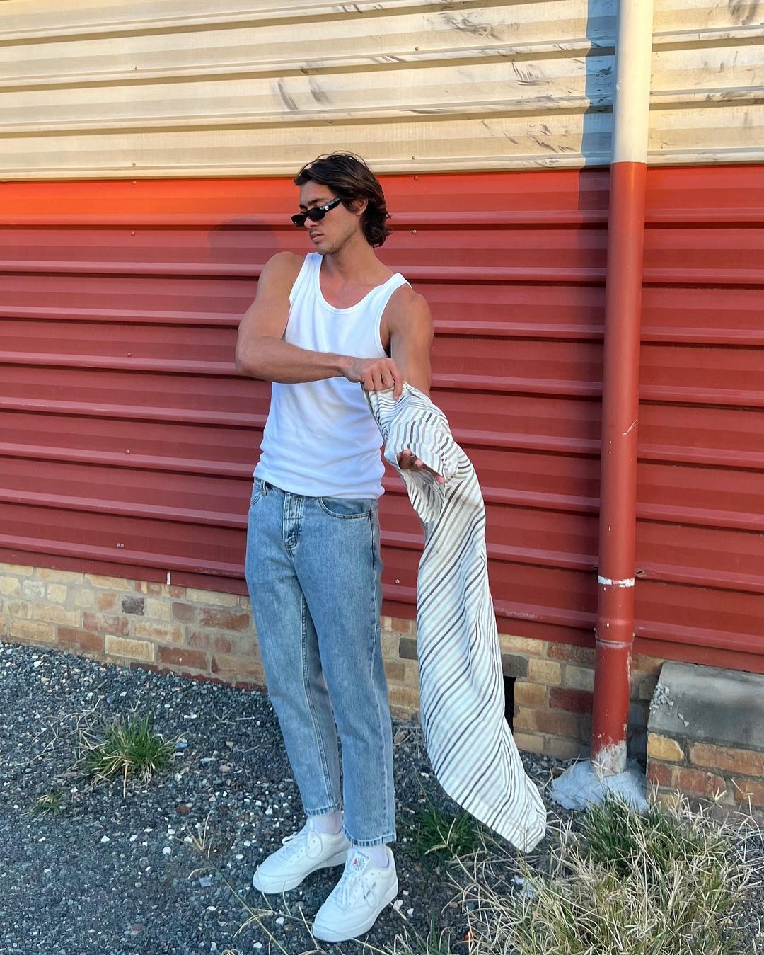 Young man poses outdoors in jeans, tank top, and sneakers, holding a striped shirt near a brick and orange wall on a sunny day.