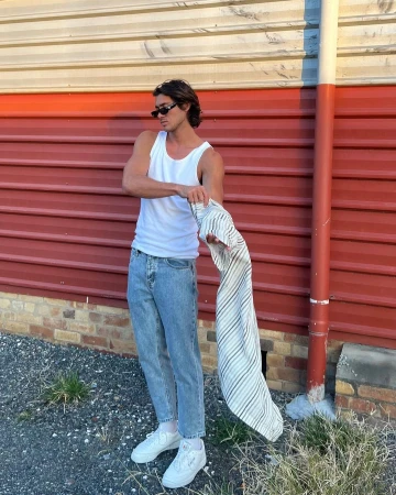 Man wearing a white tank top and blue jeans holds a striped garment against a corrugated metal wall.