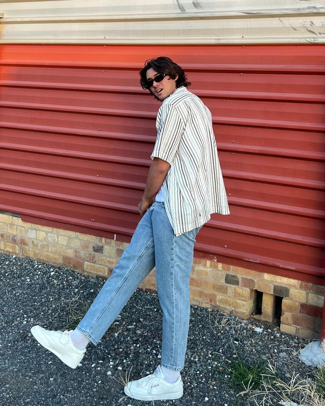 Young man poses in front of a red brick wall, wearing a vertically striped shirt, jeans, and white sneakers for a casual fashion portrait.