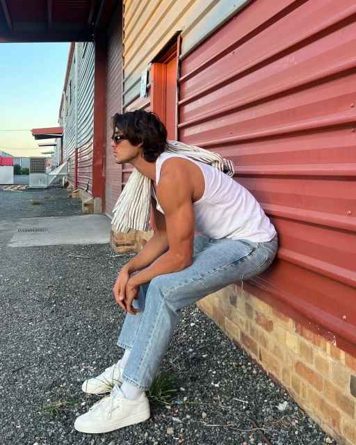 Man in tank top and jeans sits outside red building with jacket draped over shoulder. Sunglasses and white sneakers are visible.