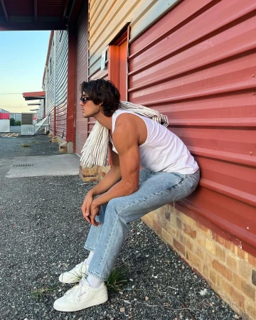 Man in tank top and jeans sits outside red building with jacket draped over shoulder. Sunglasses and white sneakers are visible.
