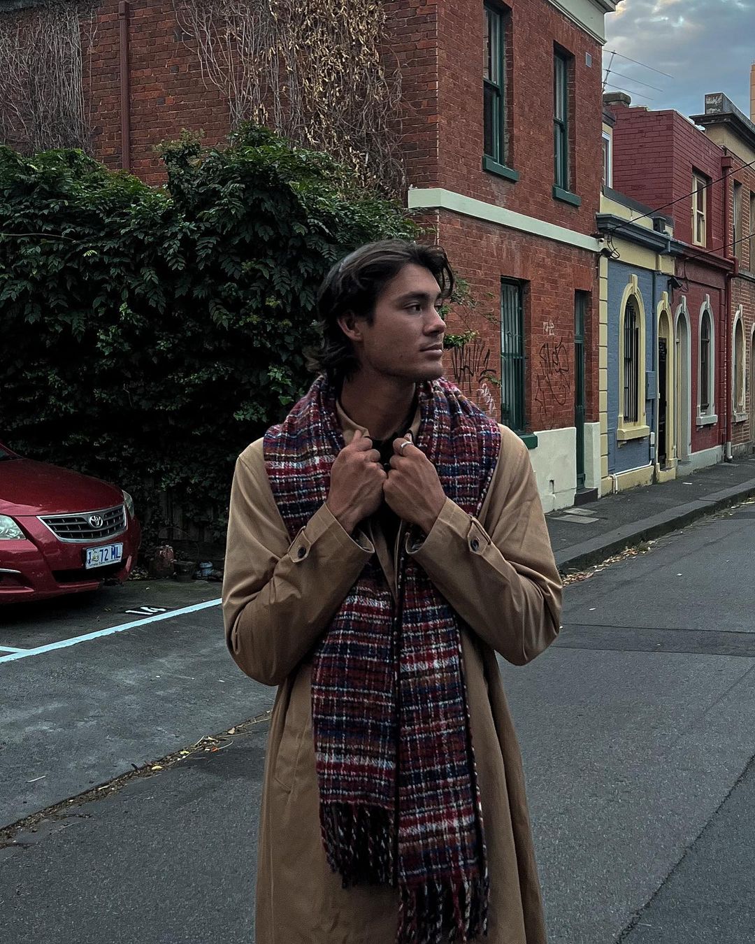 A young adult man in a trench coat and scarf stands on a city street, gazing to the side with brick buildings and a car in the background.