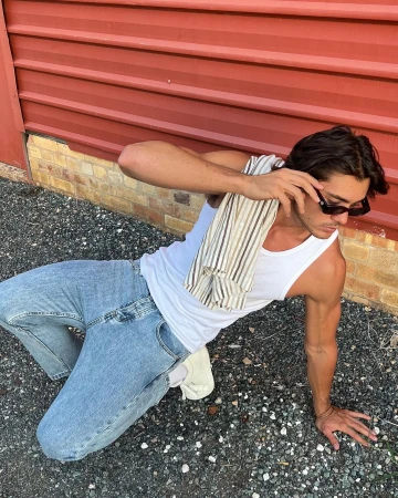 Handsome man posing outdoors in jeans and a tank top with a striped shirt draped over his shoulder, adding a casual flair.