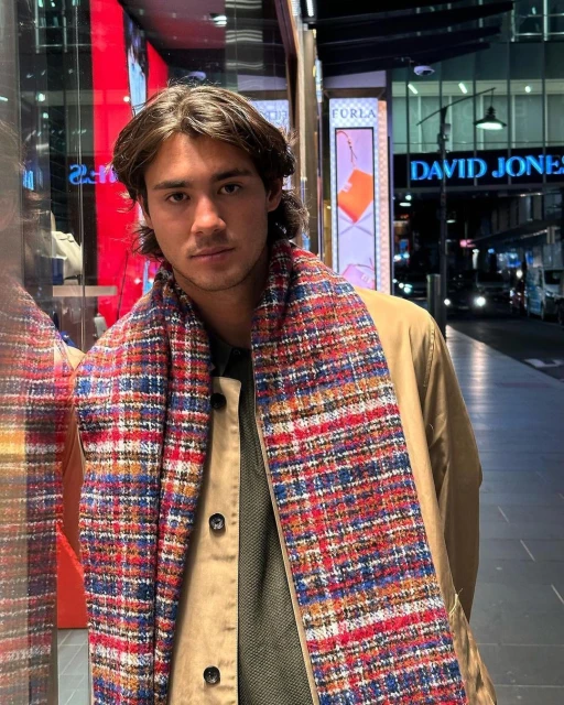 Stylish adult man wearing a multi-colored scarf and tan coat stands on a city sidewalk with store windows in the background.