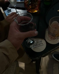 Close-up of a person holding a wine glass with dark red wine at a dark table with other glasses and a coaster.
