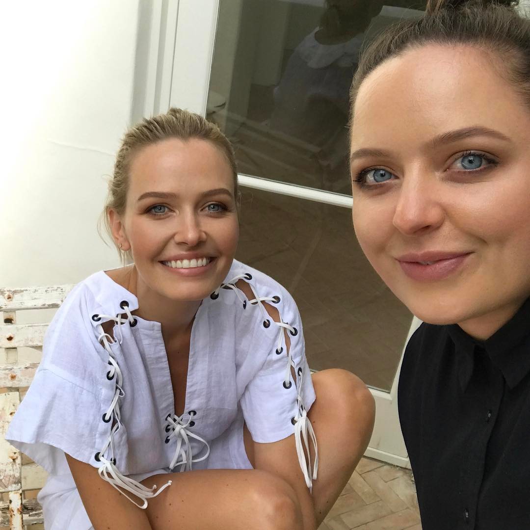 Two women pose for a selfie, one with blond hair in a white dress, the other with brown hair in a black top, smiling indoors.