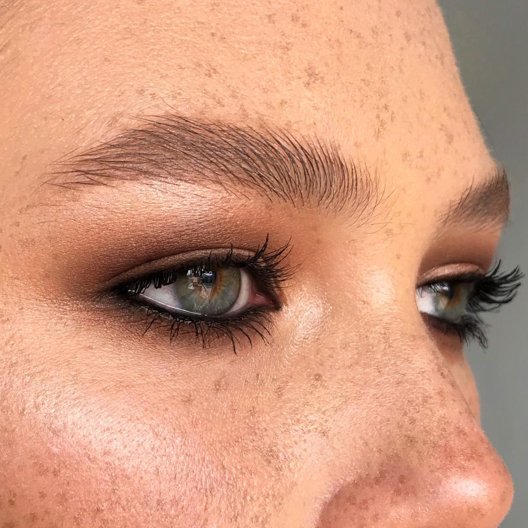 Close-up of a woman's face featuring her eyes with brown eyeshadow, black eyeliner, and full eyelashes, plus detailed freckles.