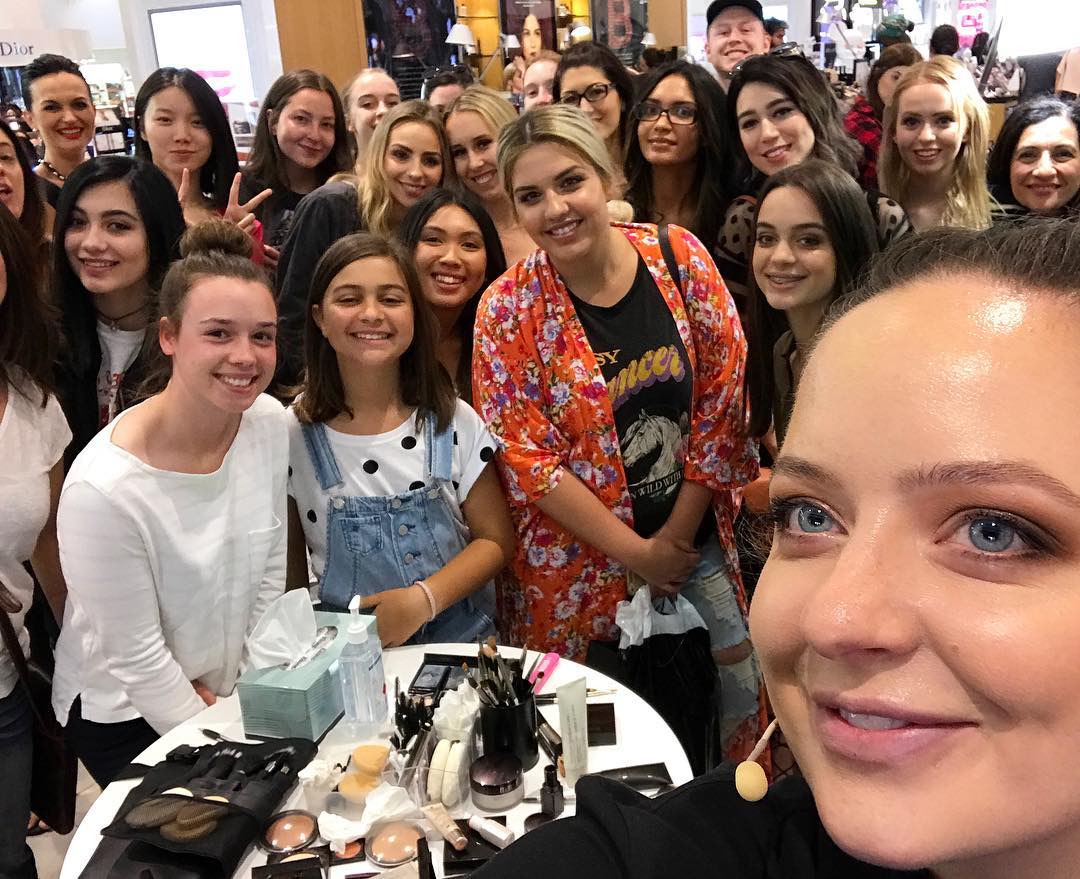 Large group of women pose for a selfie indoors with makeup, cosmetic products, and makeup brushes on a table in front of them.