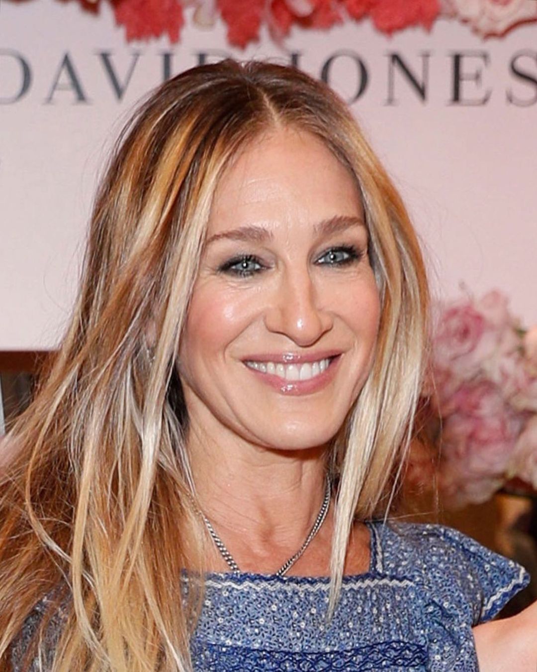 Close-up portrait of Sarah Jessica Parker, blonde hair, wearing a blue floral dress, smiling at a public event, indoor setting.