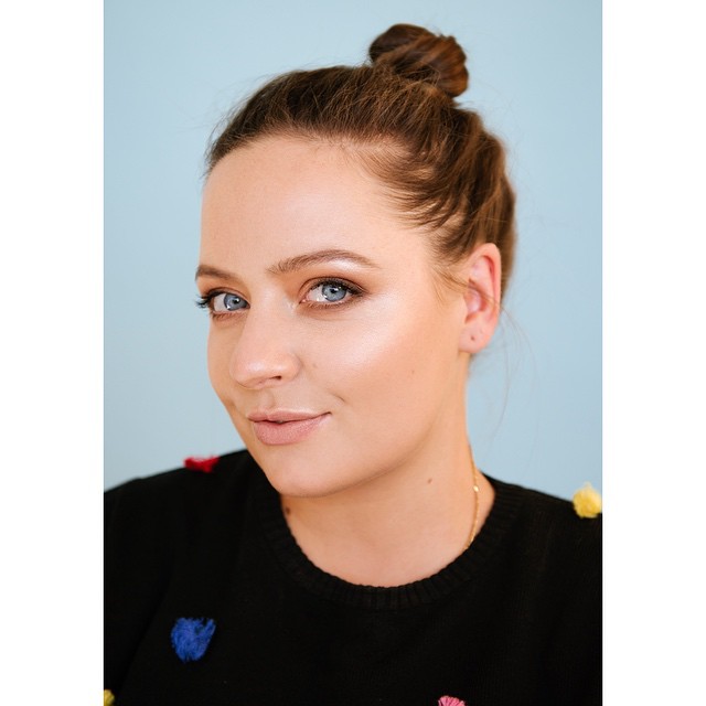 Close-up portrait of a smiling woman with a bun hairstyle, light blue eyes, and a black sweater with colorful pom-pom details.