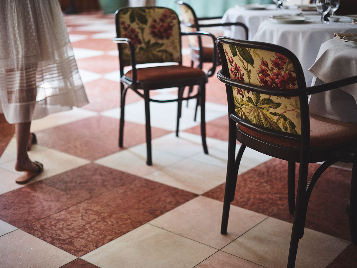 Interior shot with ornate chairs around tables with white tablecloths and a tiled floor with a woman walking past.