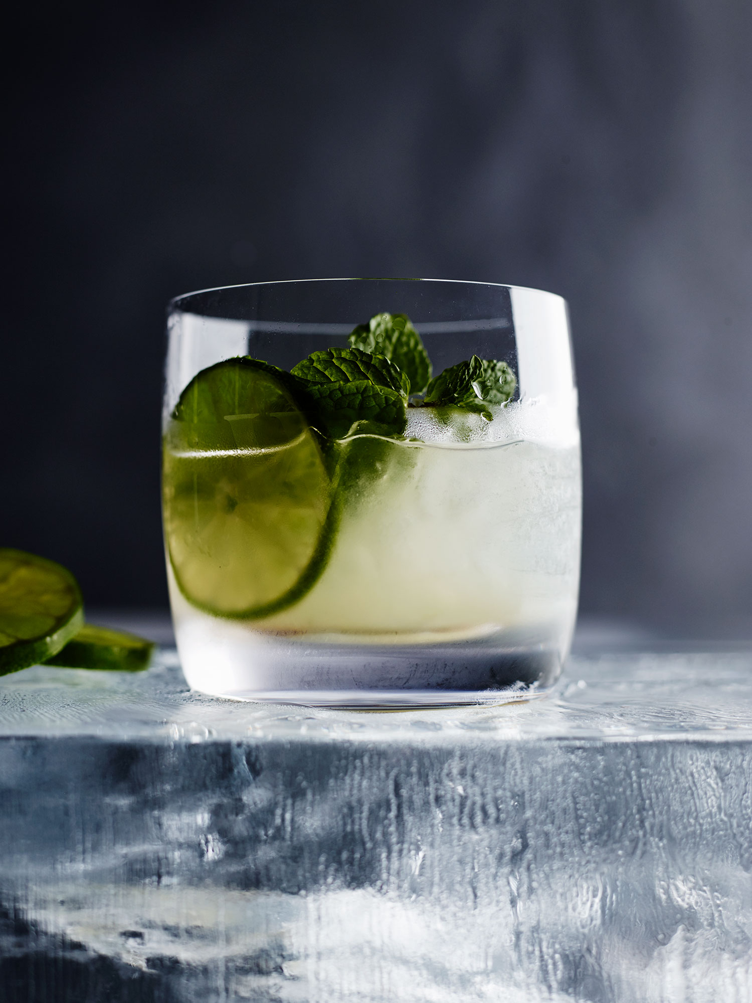 Cocktail in clear glass with ice, lime slice, and mint garnish, sitting atop textured ice block in dim lighting.