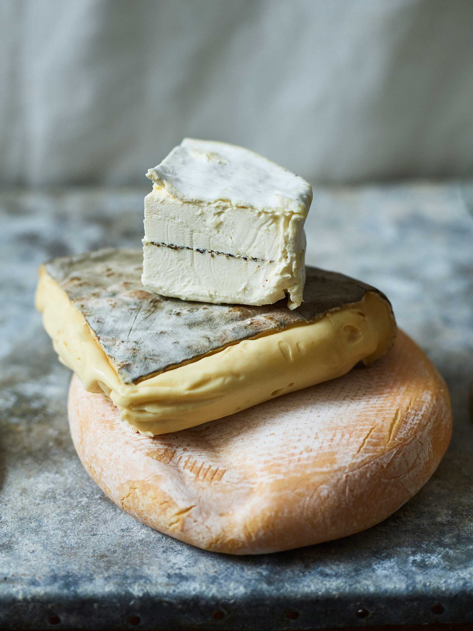 Close-up of three stacked cheese types: a white-blue block, a grey-white square, and a round orange cheese against a grey background.