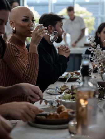 Indoor gathering shows a woman drinking, surrounded by diners at a table set with food, drinks, and decorative flowers.
