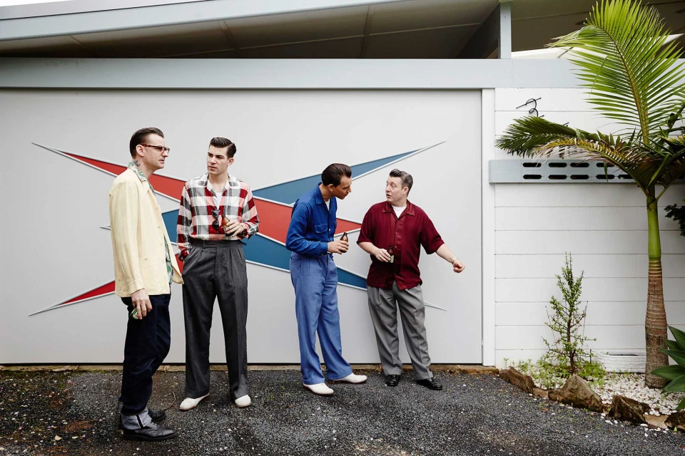 Four retro-styled men are standing outdoors in front of a building with a mid-century modern design and palm trees, casual vibe.