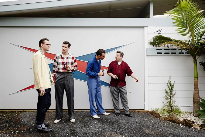 Four retro-styled men are standing outdoors in front of a building with a mid-century modern design and palm trees, casual vibe.