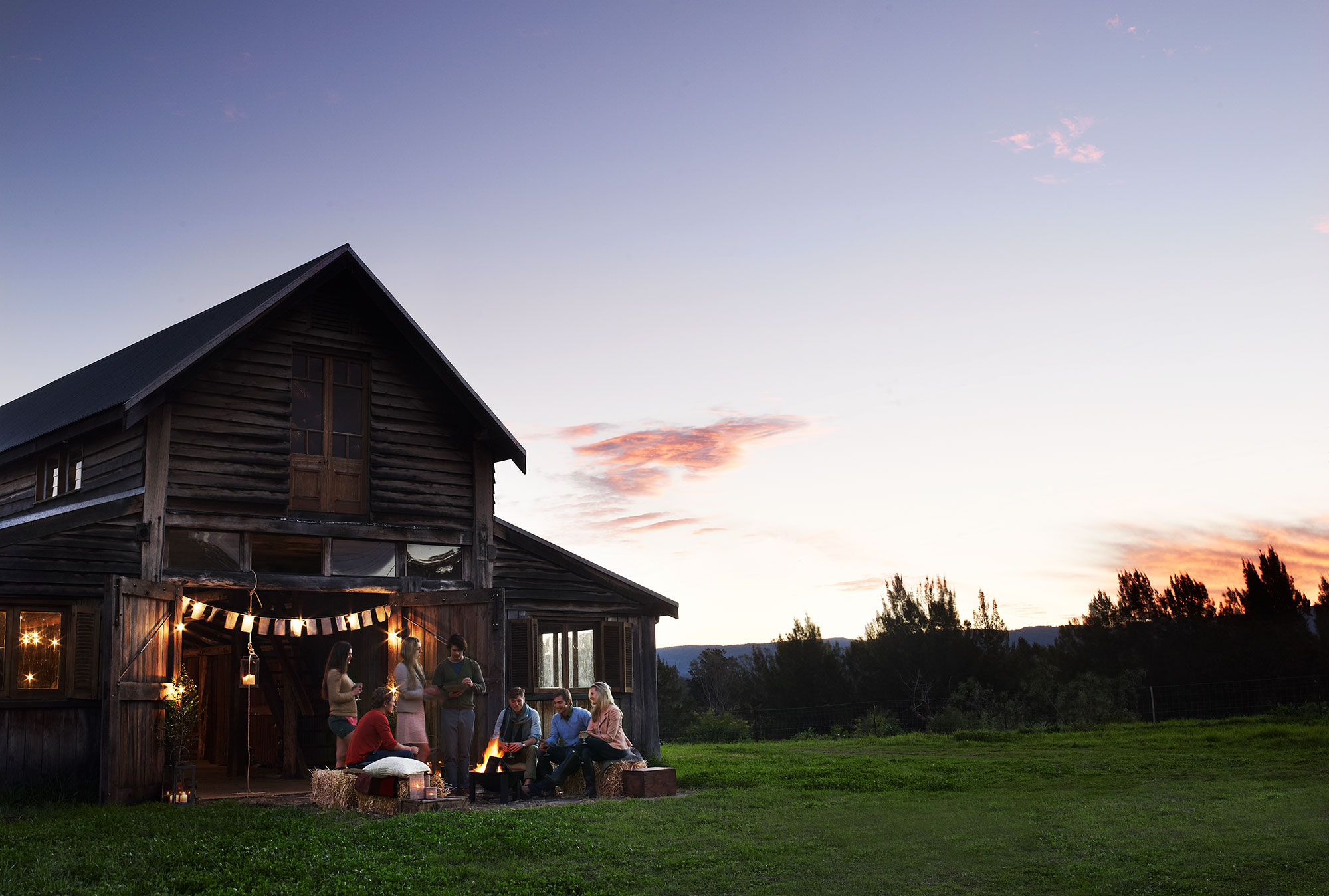 Group of people gathers outside a rustic barn at dusk with a bonfire, hay bales, and string lights creating a warm, inviting atmosphere.
