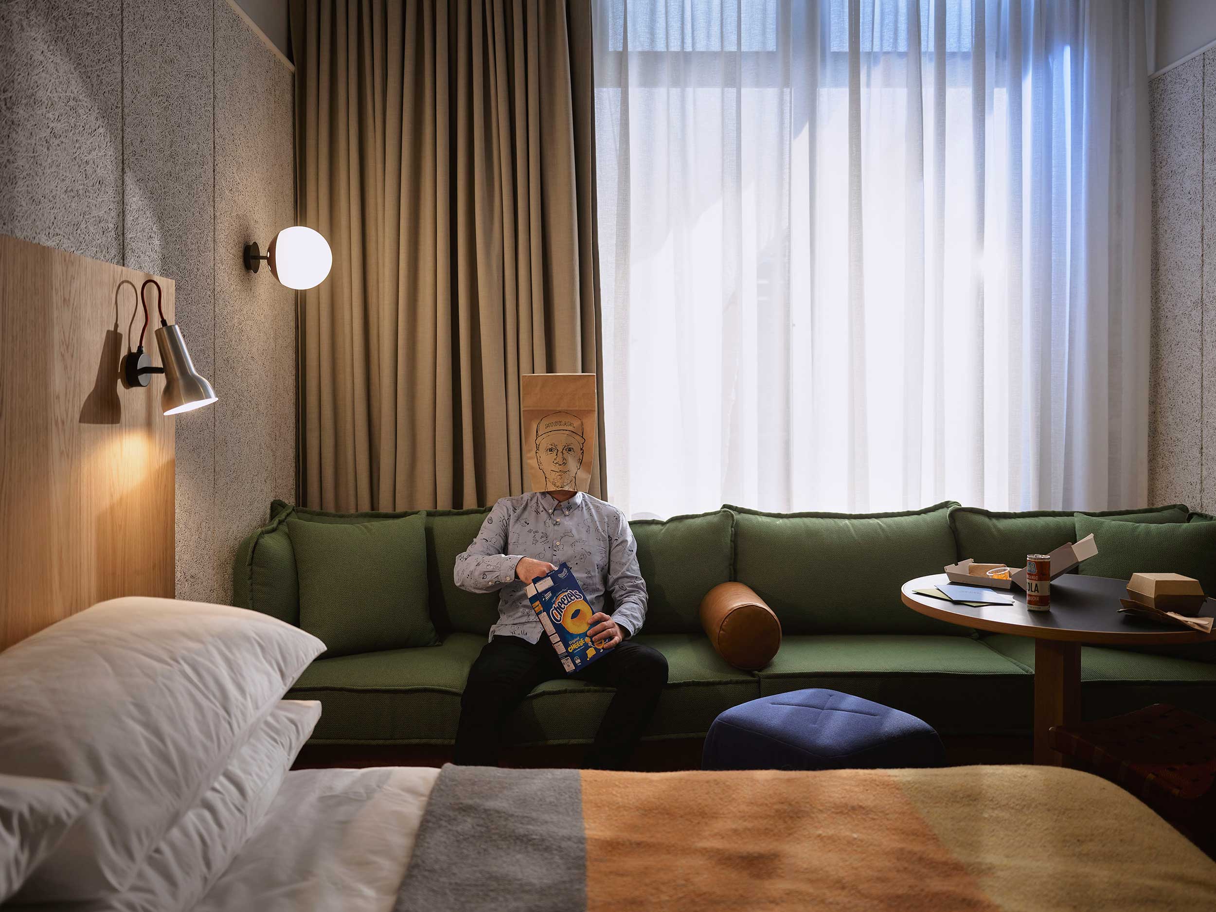Person with a box on their head sits on a green couch in an apartment next to a table with snacks and drinks by a bright window.