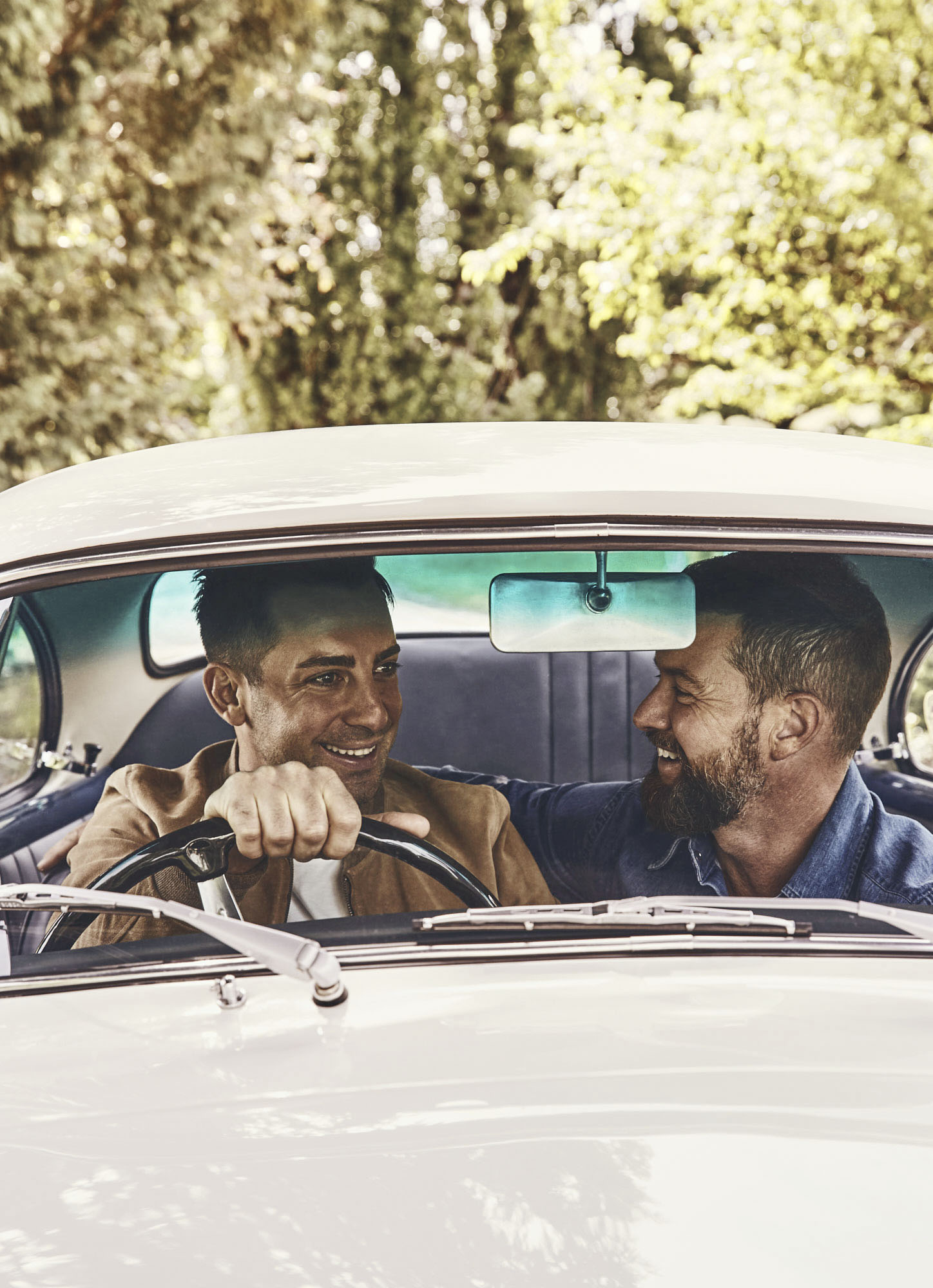 Two men are in a vintage white car, enjoying a drive on a sunny day. The driver smiles as he turns the wheel, while the passenger looks on.