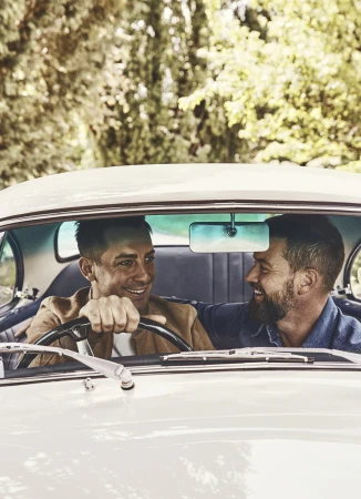 Two men are in a vintage white car, enjoying a drive on a sunny day. The driver smiles as he turns the wheel, while the passenger looks on.