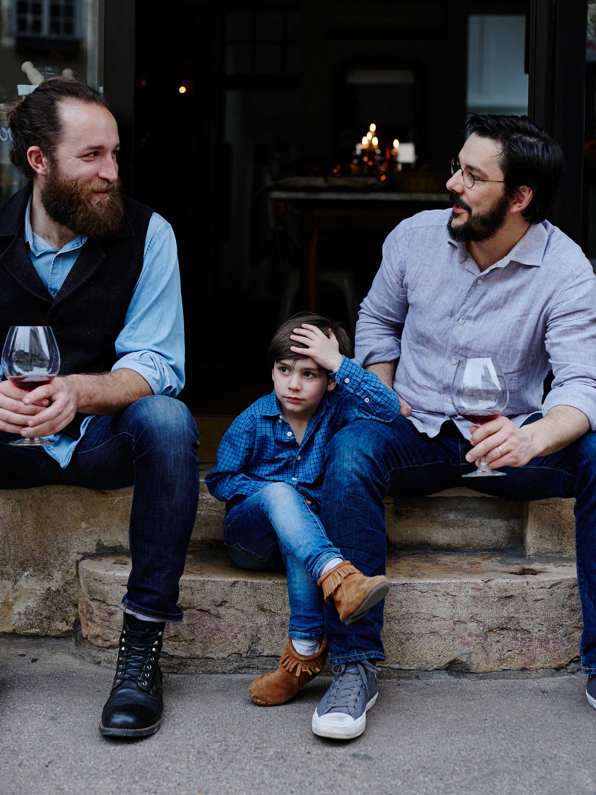 Two bearded adult men and a child sit on stone steps with wine glasses. They are outdoors, dressed casually in button-down shirts and jeans.