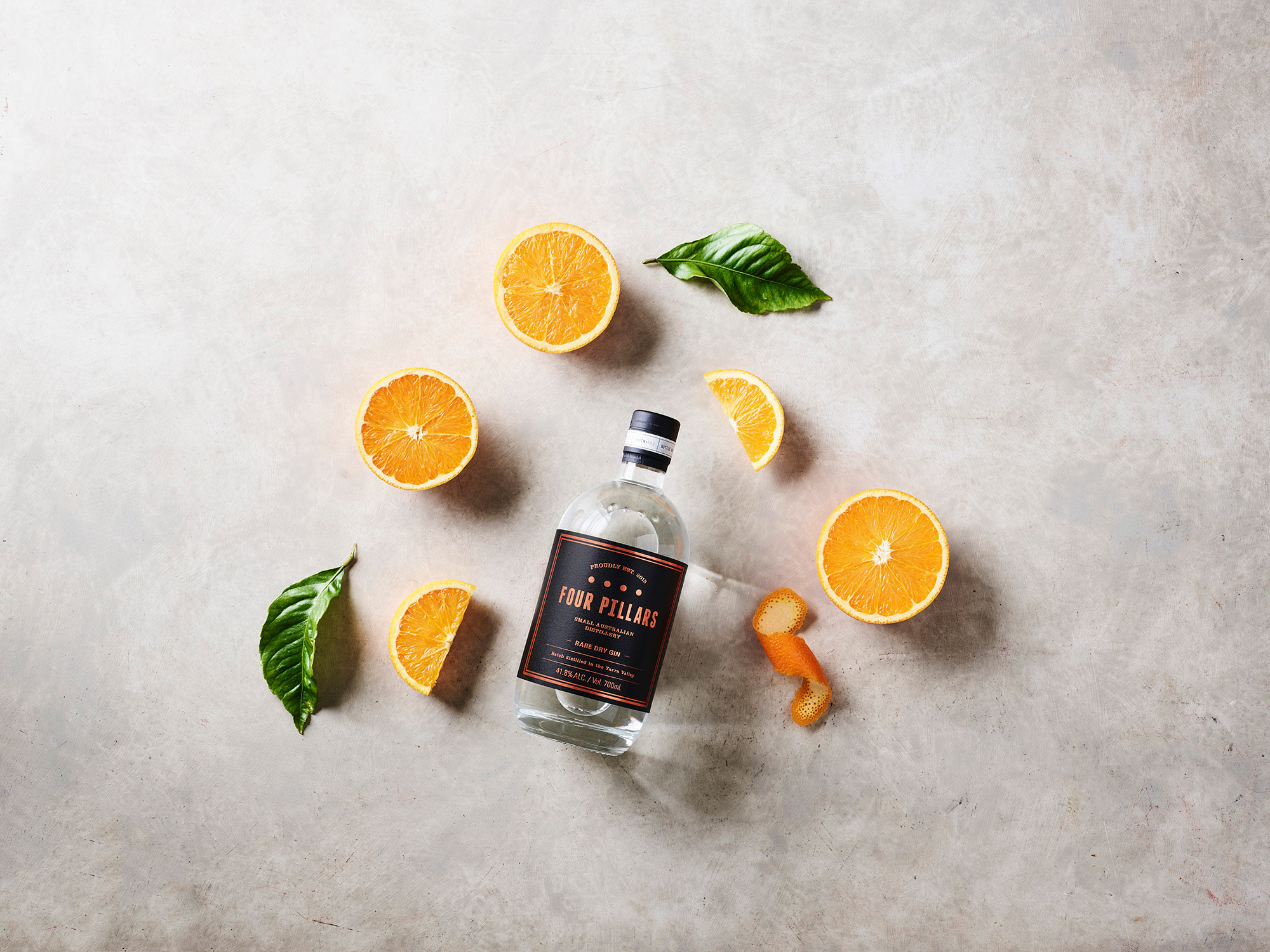 Overhead studio shot of a Four Pillars gin bottle surrounded by orange halves, orange peel, and green leaves against a light grey background.