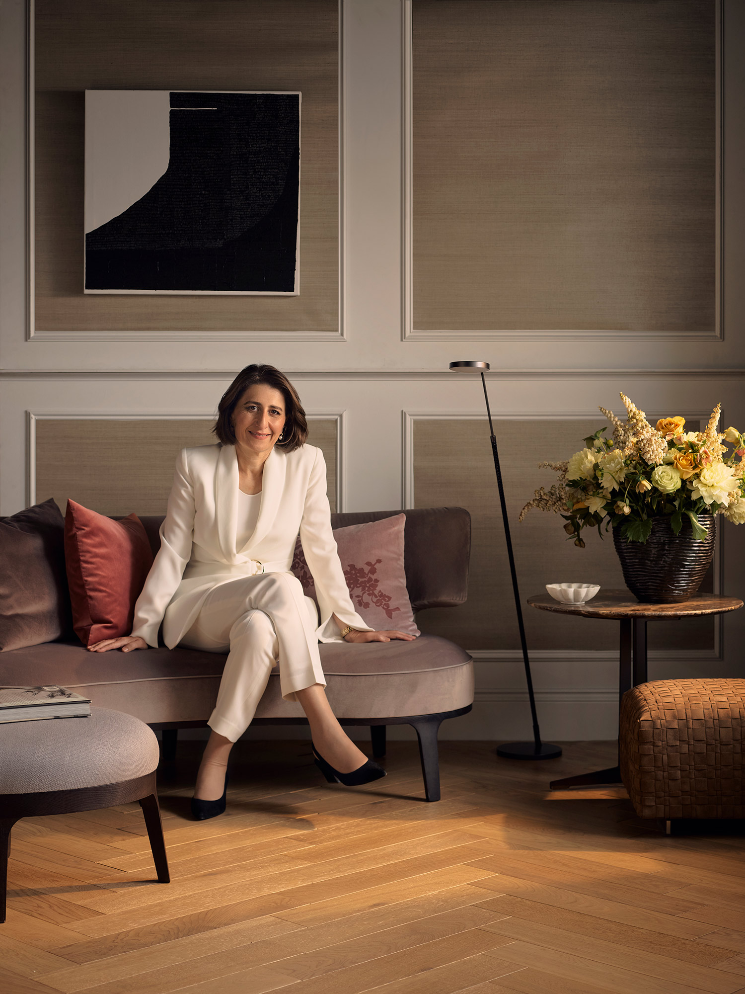 A woman in a white suit sits on a mauve couch in an elegant room, next to a side table with flowers, an abstract artwork on the wall.