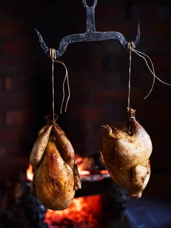 Two roasted chickens hang from a rustic metal rack in front of a brick fireplace with burning logs, creating a warm, inviting culinary scene.