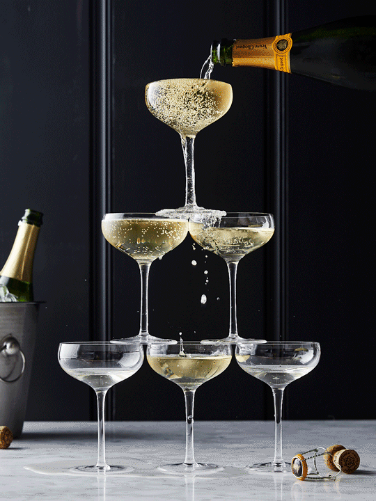 Champagne tower being filled. Six coupe glasses are stacked in a pyramid on a marble surface, and a bottle pours champagne.
