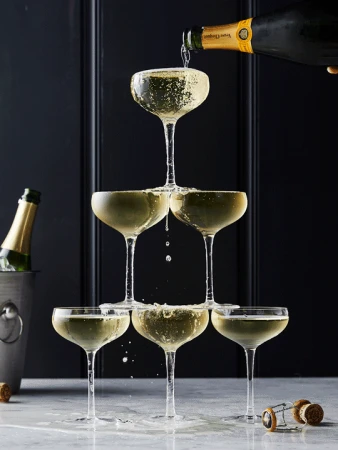 Champagne tower being filled. Six coupe glasses are stacked in a pyramid on a marble surface, and a bottle pours champagne.