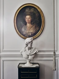 White classical marble bust on a dark cabinet below an oval framed portrait against a panelled wall