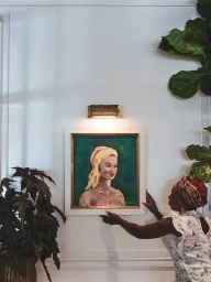 Woman in white lace top hanging a framed portrait painting on a white wall under a brass light fixture