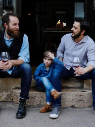 Two bearded men and a child sitting on stone steps, holding wine glasses