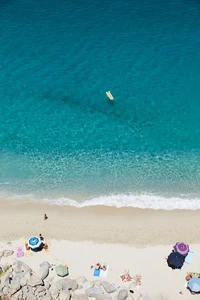 Aerial view of a beach with white sand, clear turquoise water, people sunbathing, umbrellas, and an inflatable raft floating in the ocean.