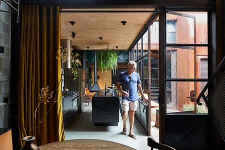 Modern home interior featuring an adult male walking through a glass doorway, showcasing a kitchen with a central island and hanging plants.