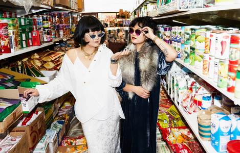 Two women pose in a brightly lit store aisle, wearing sunglasses and fashionable clothing amid shelves of products and cardboard boxes.