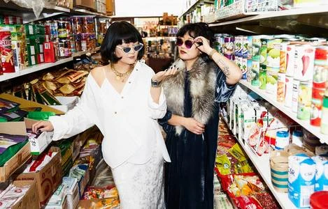 Two women pose in a brightly lit store aisle, wearing sunglasses and fashionable clothing amid shelves of products and cardboard boxes.