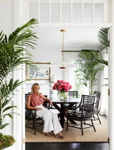 Blonde woman in a mauve top and white pants sits at a round dining table adorned with pink peonies, surrounded by tropical plants.