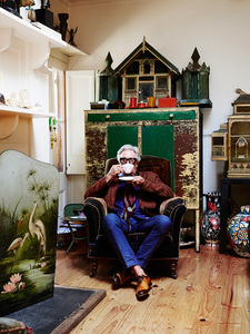 Indoor portrait of an elderly man in a brown jacket, blue jeans, and glasses, sitting in an armchair and drinking tea.