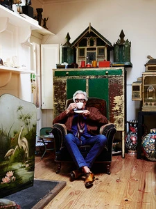 Indoor portrait of an elderly man in a brown jacket, blue jeans, and glasses, sitting in an armchair and drinking tea.