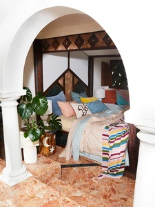 View of a bedroom with a four-poster bed, colorful bedding, and plants in decorative vases, framed by an arched opening and tiled floor.