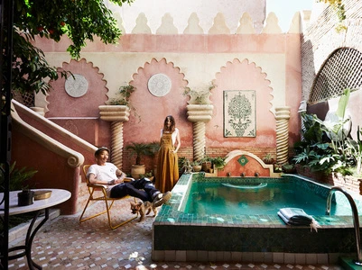 A couple poses in the courtyard of a riad with a small pool. The man sits in a yellow chair as the woman stands next to the pool.
