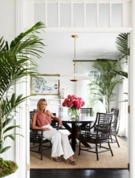 Woman seated in a dining chair inside a bright room decorated with lush potted plants and a dining table with pink flowers.
